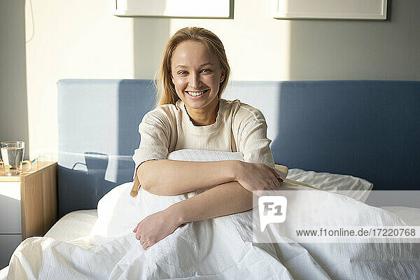 Junge Frau lächelt  während sie die Knie auf dem Bett zu Hause umarmt