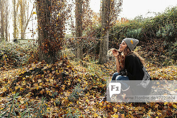 Lächelnde Frau mit Hand im Haar  die im Herbstwald sitzt und nachdenkt