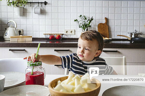 Niedlicher Junge hält Strohhalm in Einmachglas  während er am Esstisch in der Küche sitzt