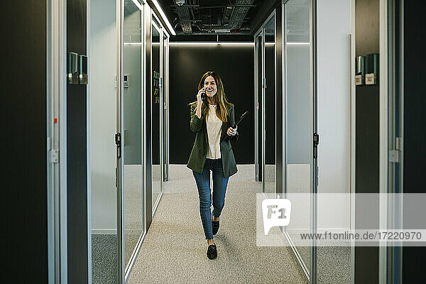 Selbstbewusste Geschäftsfrau  die auf einem Smartphone spricht  während sie im Büro durch den Korridor geht