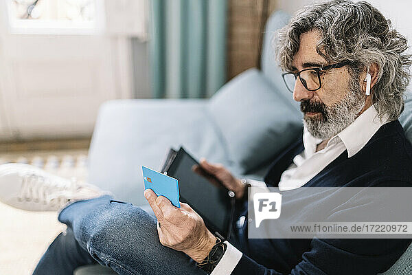 Handsome man looking at credit card holding digital tablet while sitting at home