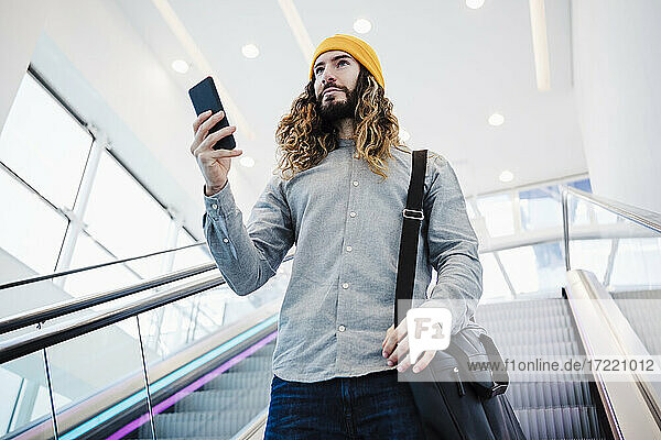 Man with long hair holding mobile phone while moving on escalator