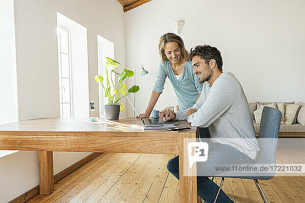 Schöne Frau  die einen Mann ansieht  der an einem Laptop arbeitet  während er auf einem Schreibtisch im Büro zu Hause sitzt