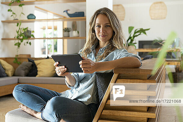 Glückliche Frau  die ein Tablet hält und mit gekreuzten Beinen auf der Couch im Wohnzimmer sitzt