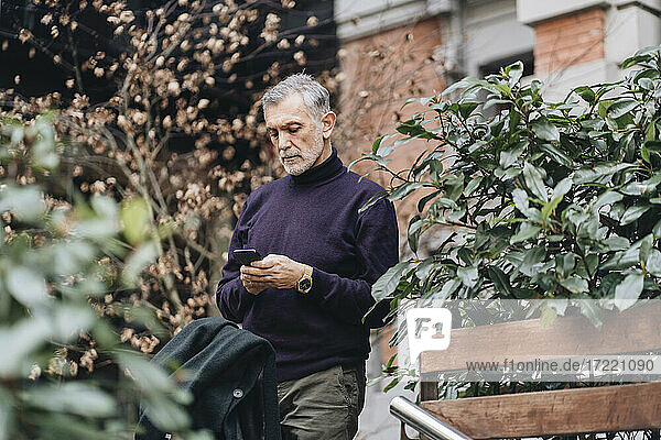Mature man using mobile phone while standing by plant