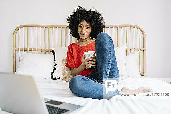 Frau hält Tasse und schaut auf den Laptop  während sie zu Hause auf dem Bett sitzt