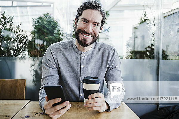 Glücklicher männlicher Geschäftsmann mit Smartphone und wiederverwendbarem Kaffeebecher im Büro