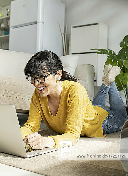 Fröhliche Frau mit Brille benutzt einen Laptop  während sie zu Hause auf dem Boden liegt