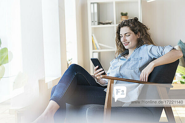 Lächelnde Frau  die ein Smartphone benutzt  während sie sich auf einem Stuhl zu Hause entspannt