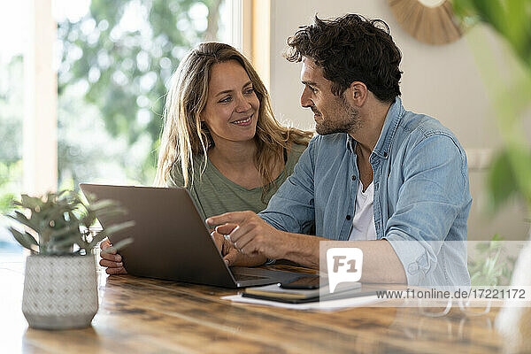 Smiling couple looking at each other while sitting in front of laptop at table