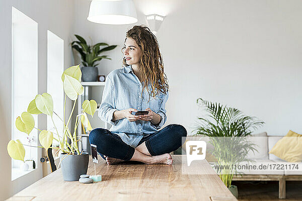 Smiling woman with brown hair looking away while holding mobile phone on table at home