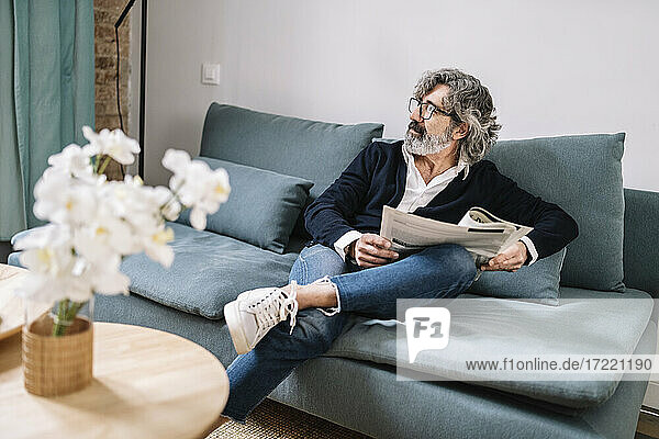 Hübscher älterer Mann mit Zeitung  der wegschaut  während er zu Hause auf dem Sofa sitzt