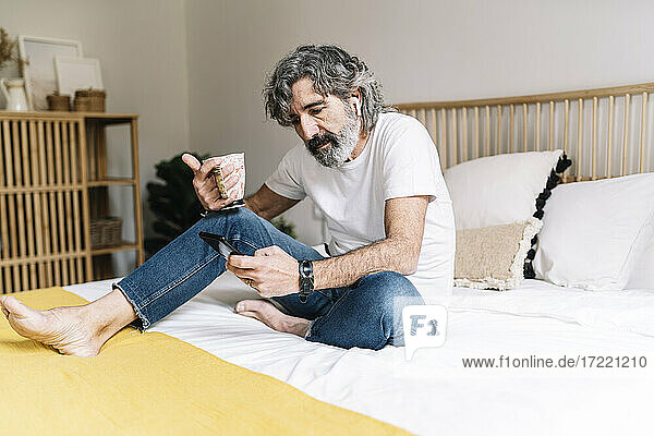 Mann  der ein Smartphone benutzt und einen Kaffeebecher hält  während er zu Hause auf dem Bett sitzt