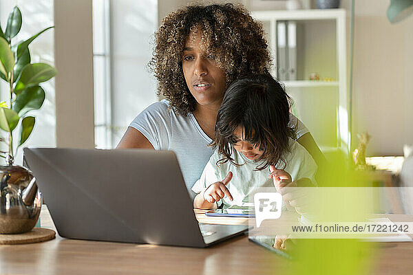 Mother working on laptop while her daughter playing with smart phone in home office