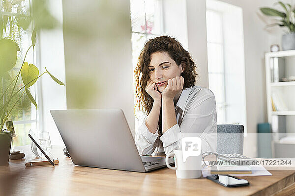 Geschäftsfrau mit Hand am Kinn,  die auf einen Laptop im Heimbüro schaut