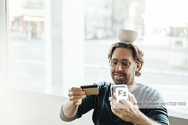 Man balancing bowl on head while paying through credit card on smart phone in coffee shop