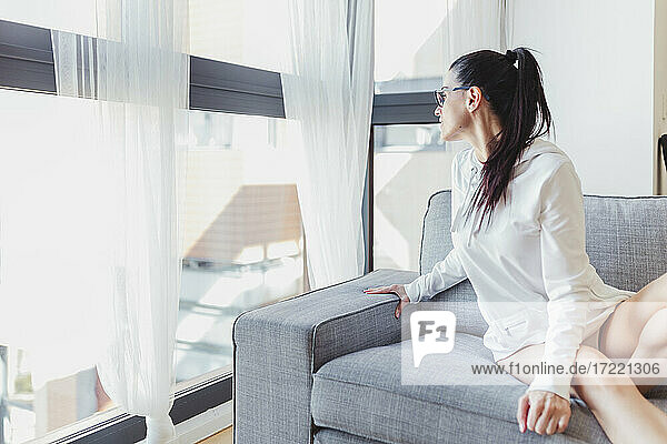 Frau schaut durch das Fenster  während sie auf dem Sofa im Wohnzimmer sitzt