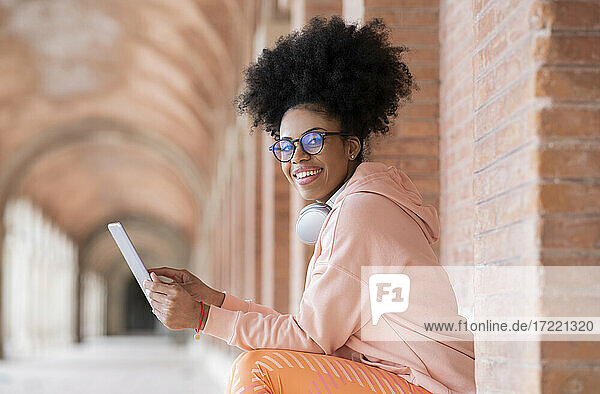 Lächelnde Frau mit Brille  die ein digitales Tablet benutzt  während sie an einer Wand im Korridor sitzt