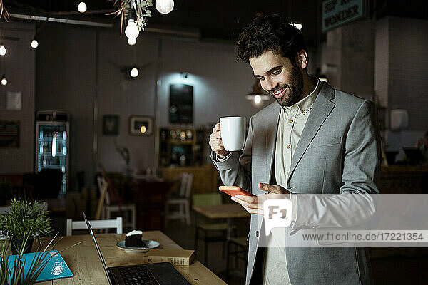 Lächelnder männlicher Unternehmer  der ein Smartphone benutzt  während er einen Kaffee in einem Café trinkt