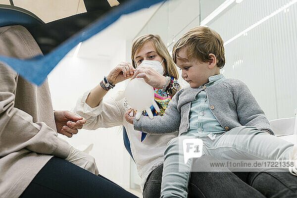 Junge spielt mit aufblasbarem Schutzhandschuh  während er mit seiner Mutter beim Zahnarzt in der Klinik sitzt