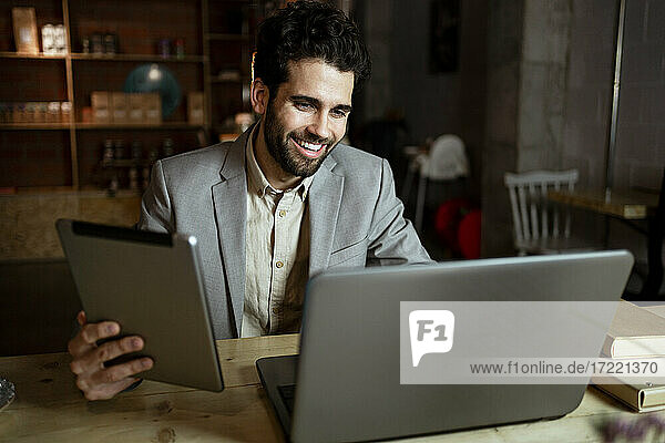 Glücklicher männlicher Freiberufler mit digitalem Tablet bei einem Videogespräch über einen Laptop in einem Café
