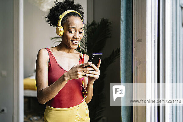 Junge Frau mit Kopfhörern  die ein Smartphone benutzt  während sie zu Hause am Fenster steht