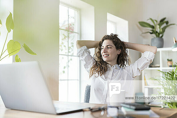 Lächelnde weibliche Fachkraft mit Händen hinter dem Kopf  die sich am Schreibtisch im Heimbüro entspannt