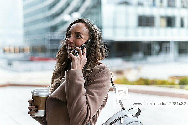 Glückliche Geschäftsfrau  die einen Einweg-Kaffeebecher hält  während sie mit einem Mobiltelefon spricht