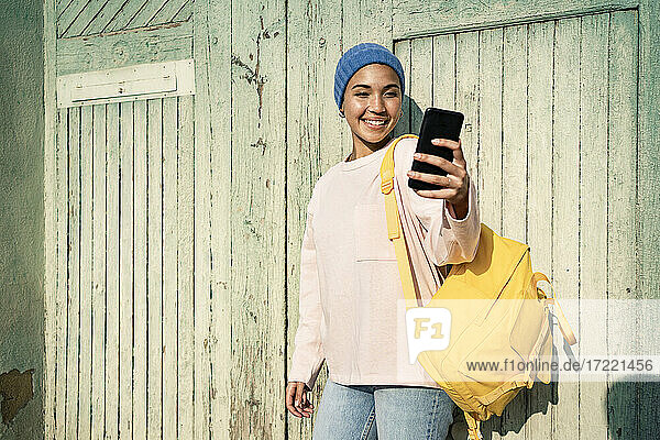 Lächelnde Frau mit Rucksack nimmt Selfie durch Smartphone während sonnigen Tag