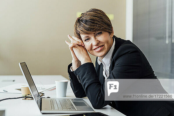 Glückliche Geschäftsfrau mit verschränkten Händen vor einem Laptop am Schreibtisch im Büro sitzend