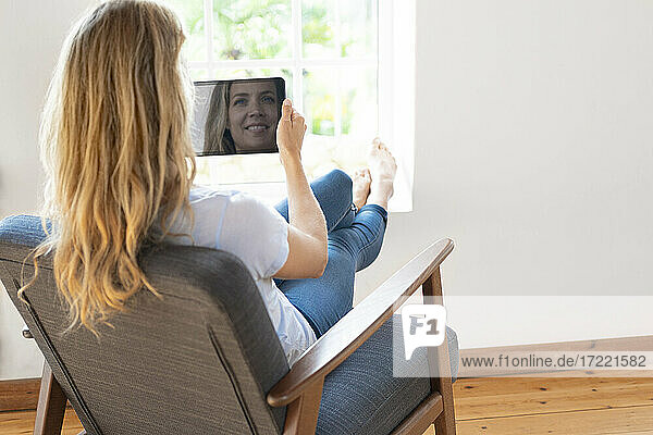 Blond woman holding digital tablet while relaxing on chair at home