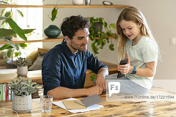 Mädchen zeigt ihrem Vater das Handy  während sie zu Hause auf dem Tisch sitzt