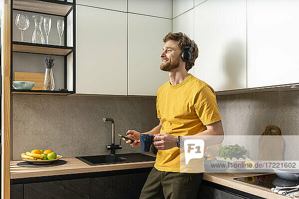 Lächelnder Mann hält Kaffeetasse und Smartphone  während er in der Küche über Kopfhörer Musik hört