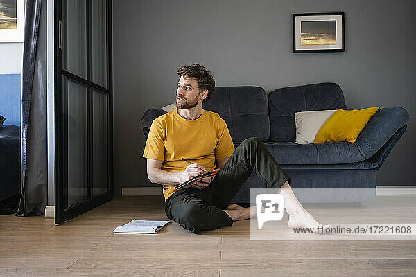 Nachdenklicher Mann hält ein Buch in der Hand und sitzt auf dem Boden im Wohnzimmer