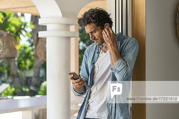 Mann  der ein Smartphone benutzt  während er über Kopfhörer am Türrahmen Musik hört