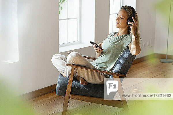 Frau hört Musik über Kopfhörer und hält ihr Handy auf einem Sessel im Wohnzimmer