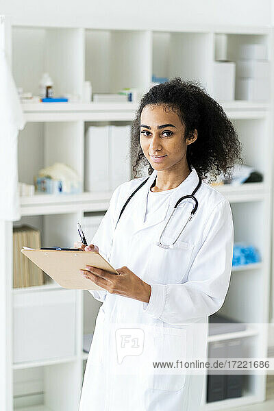 Lächelnder junger Arzt mit Klemmbrett und Stift in der Hand  während er in einer Klinik steht