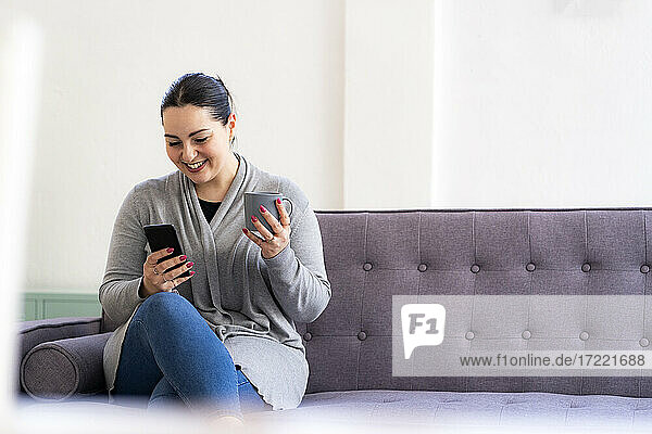 Lächelnde Frau  die ein Mobiltelefon benutzt und eine Kaffeetasse zu Hause hält