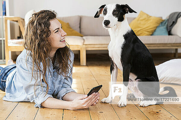Schöne Frau mit langen Haaren  die ihr Smartphone hält und einen Hund im Wohnzimmer betrachtet