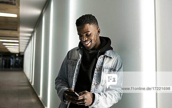 Lächelnder junger Mann  der ein Smartphone benutzt  während er an der Wand steht