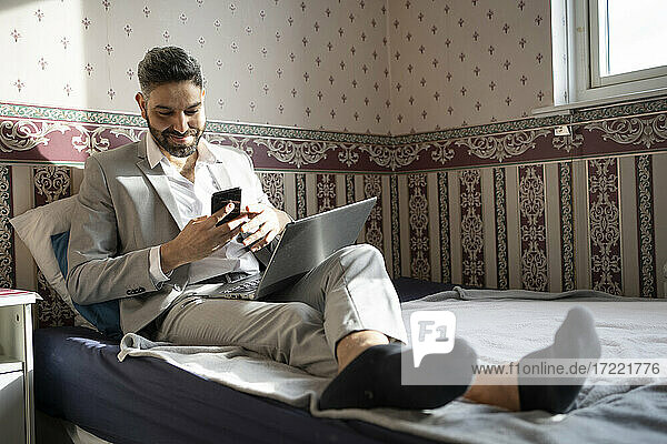 Lächelnder männlicher Fachmann  der ein Smartphone benutzt  während er zu Hause auf dem Bett sitzt