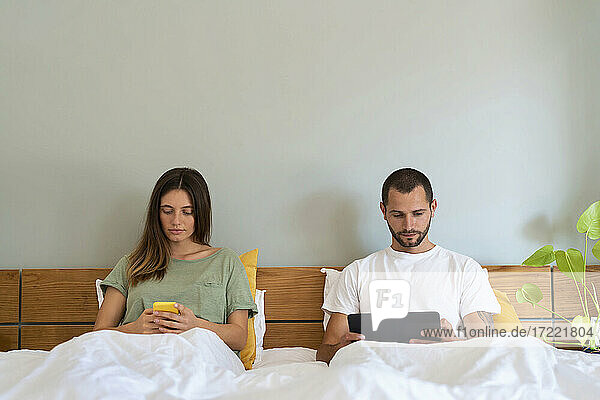 Junges Paar benutzt Smartphone und Tablet  während es zu Hause auf dem Bett sitzt