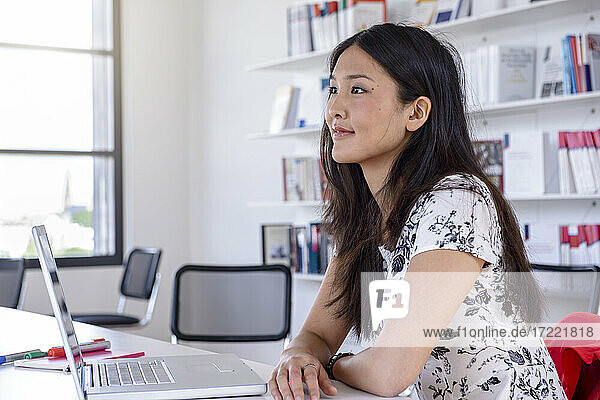 Nachdenkliche junge Frau sitzt mit Laptop am Schreibtisch in einer Bibliothek