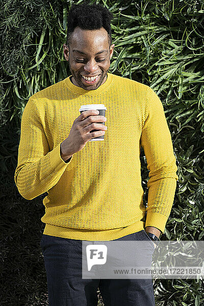 Smiling young man looking at disposable coffee cup in front of lush foliage