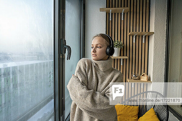 Junge Frau hört Musik über Kopfhörer  während sie mit verschränkten Armen am Fenster steht