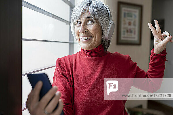Glückliche Frau gestikuliert  während sie ein Mobiltelefon im Wohnzimmer hält