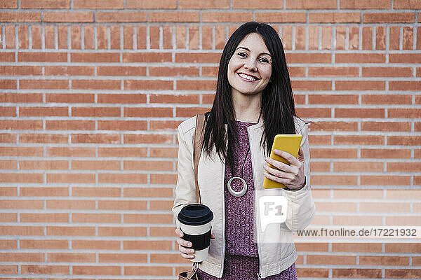 Lächelnde schöne Frau hält Smartphone und Einweg-Kaffeebecher vor einer Backsteinmauer