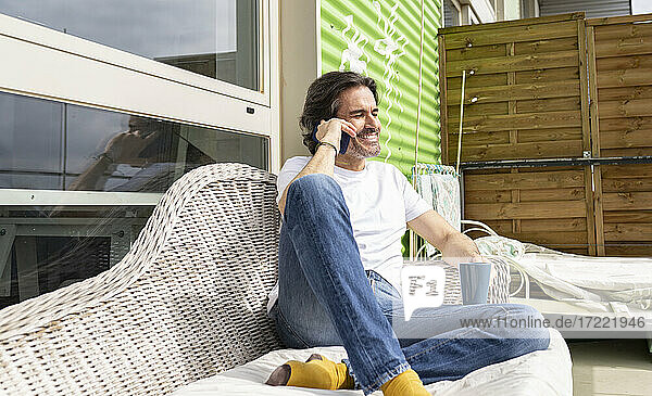 Smiling man talking on smart phone while sitting on sofa in balcony