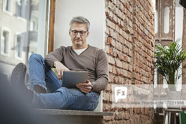 Mature man in eyeglasses using digital tablet while sitting on window sill at home