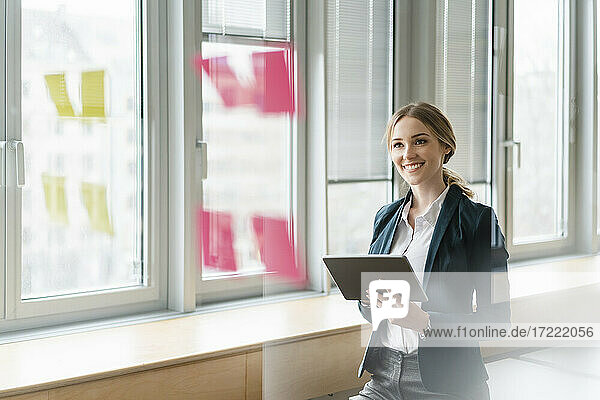 Zufriedene Geschäftsfrau mit digitalem Tablet und Blick auf Haftnotizen  die auf einem Glas im Büro kleben